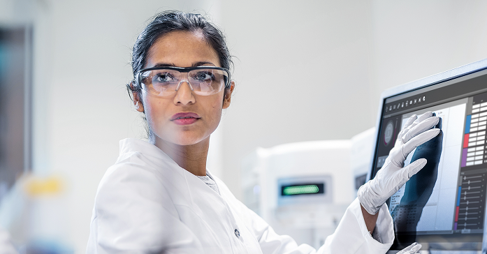 Científica con gafas de seguridad y bata blanca operando una pantalla táctil digital en un laboratorio.