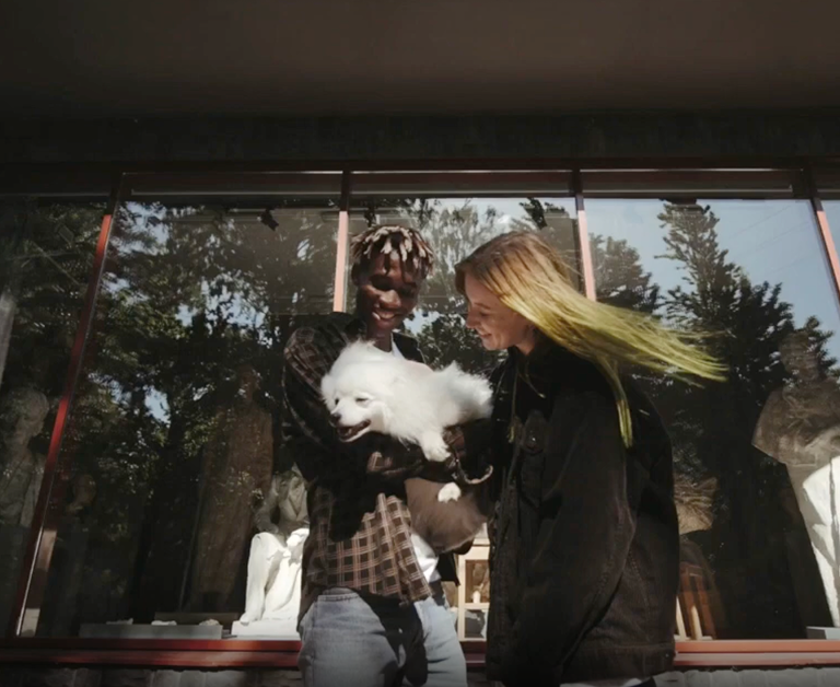 Pareja con perro blanco Un hombre y una mujer sonriendo mientras sostienen a un perro blanco pequeño frente a un gran ventanal con reflejos de árboles.