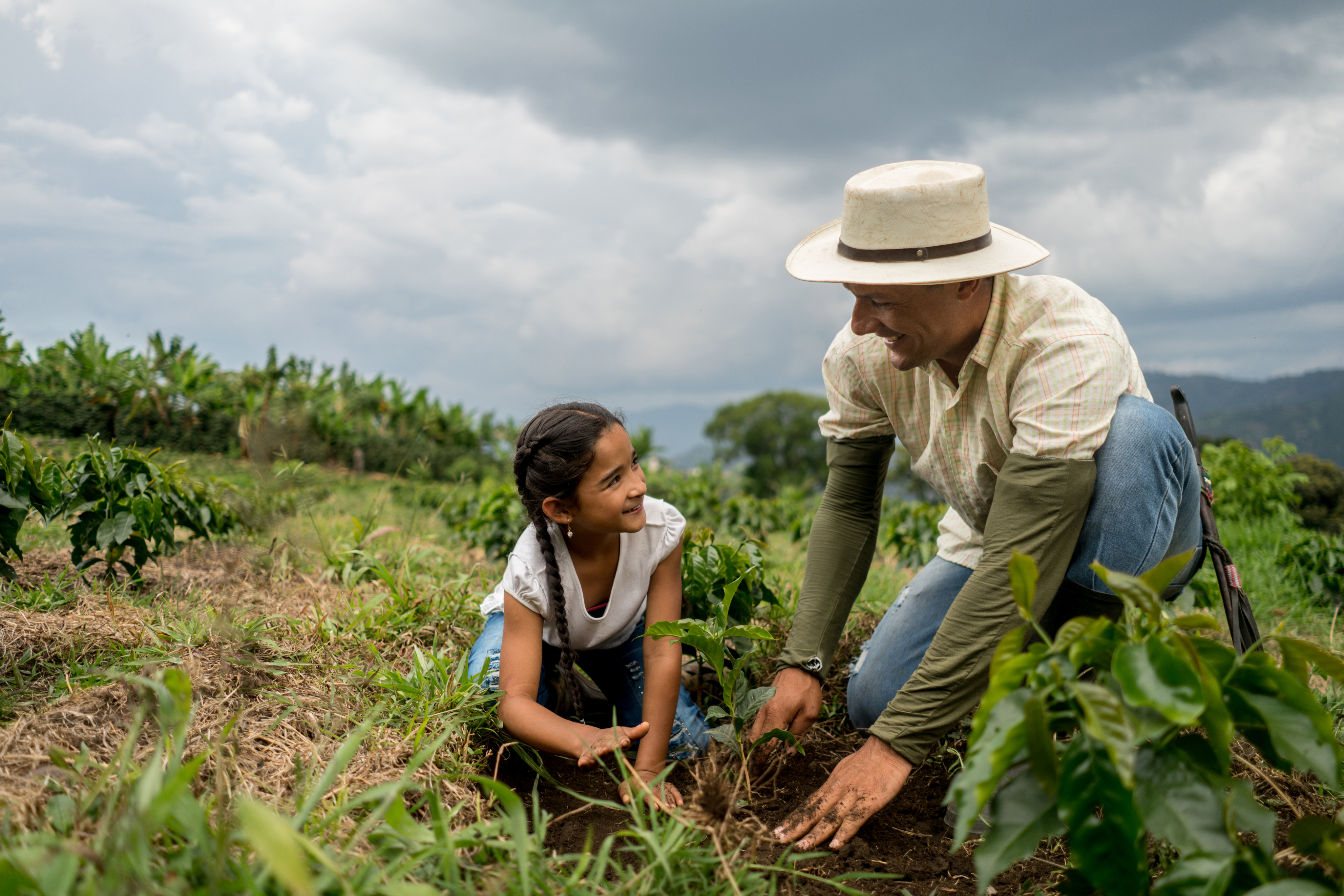Un hombre con sombrero y una niña plantando un brote en un campo bajo un cielo nublado.