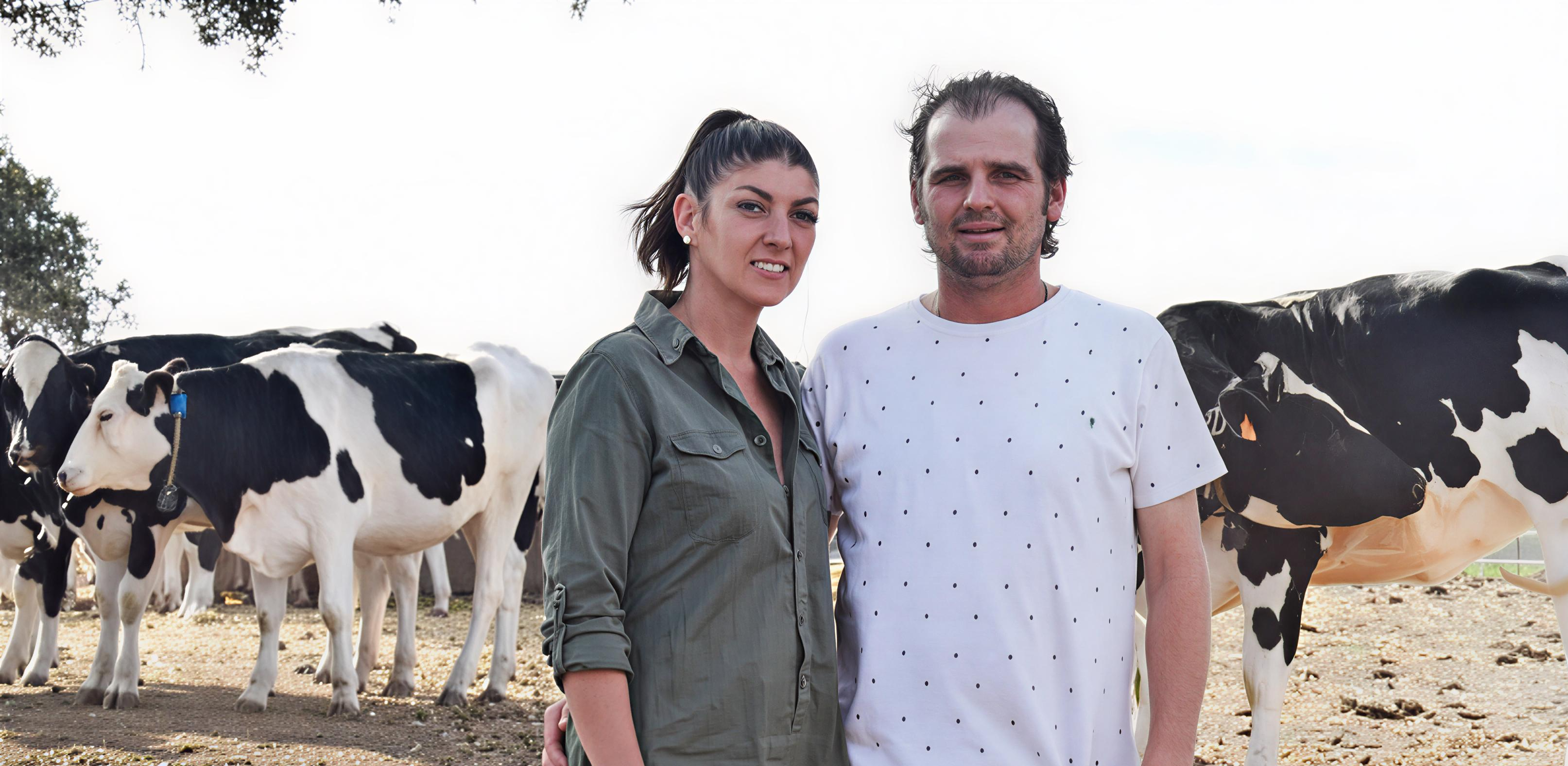 Una mujer y un hombre posando juntos en primer plano con vacas Holstein en el fondo de una granja.